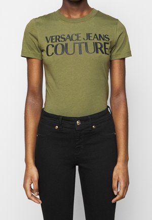 Persoon draagt een olijfgroene t-shirt met de tekst "VERSACE JEANS COUTURE" en een hooggesneden zwarte jeans, staand tegen een witte achtergrond.