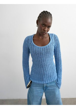 Femme aux cheveux tressés portant un pull bleu clair en crochet à manches longues et un jean, debout contre un mur blanc uni.