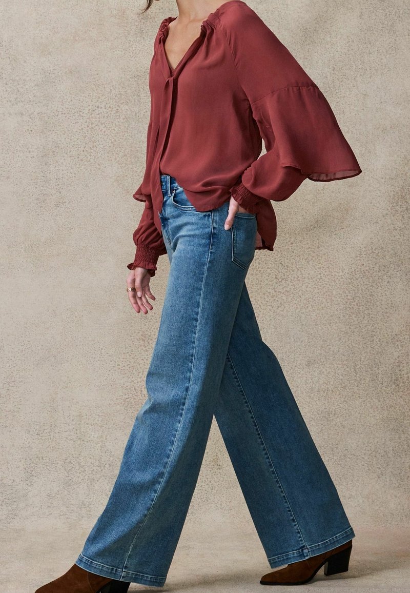 Femme portant un chemisier bordeaux ample et un jean bleu large avec des bottines marron à talons, debout devant un fond texturé uni.