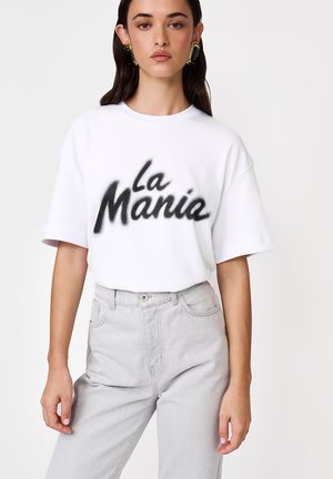 Kobieta w białym, oversize t-shircie z napisem "la Mania" czarnym ozdobnym pismem oraz jasnoszarych, wysokich jeansach na jednolitym tle.