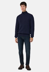 Temno moder pulover z pletenim vzorcem in visokim ovratnikom, v kombinaciji s temno modrimi kavbojkami in rjavimi čipkastimi čevlji. Pulover ima teksturirane vzorce.