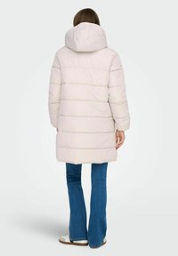 Frau steht mit dem Rücken zur Kamera, trägt einen hellbeigen gepolsterten Kapuzenmantel, blaue Jeans und weiße Turnschuhe vor einem schlichten Hintergrund.