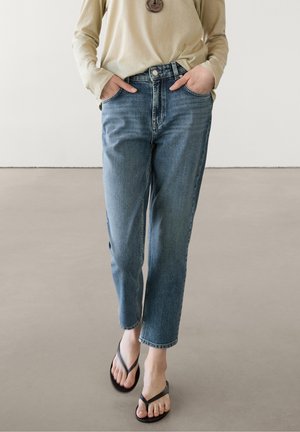 Persoon draagt blauwe recht gesneden jeans, beige top met lange mouwen en zwarte slippers, met handen in de zakken, staand op een gladde vloer.