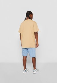 Tricou oversized din bumbac bej, cu mâneci scurte, asortat cu pantaloni scurți din denim albastru deschis și pantofi sport albi. Design simplu, croială lejeră.