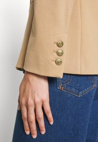 Main reposant sur le côté, portant un jean bleu et une manche de blazer beige avec trois boutons dorés décoratifs.
