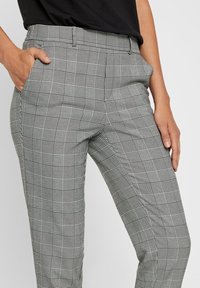 Person klädd i grå och svart rutiga skräddarsydda byxor med fickor, ihop med en svart skjorta som är instoppad i midjan.