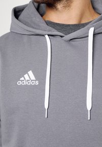 Sweat à capuche Adidas gris avec cordons blancs porté par-dessus un t-shirt gris foncé, affichant le logo de la marque sur la poitrine.