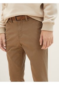 Muž oblečený v světle béžovém sv sweateru a hnědých kalhotách s odpovídajícím hnědým semišovým páskem, stojící s uvolněnýma rukama podél těla.