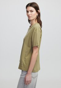 Camiseta de manga corta en verde olivo con adornos de tachuelas plateadas en el frente. Hecha de una tela suave con un corte relajado.