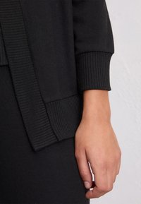 Mano appoggiata accanto a un maglione con polsini a coste neri e orlo coordinato su uno sfondo chiaro e semplice.