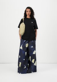 Camiseta negra de manga corta con un pequeño logo blanco, combinada con pantalones de pierna ancha estampados en azul oscuro y crema. Mochila de cuerda beige.