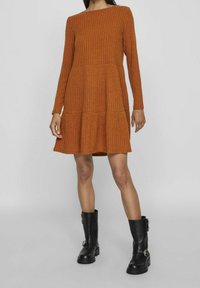 Femme portant une robe longue à manches longues côtelée de couleur rouille, à volants, avec des bottes noires à boucle mi-mollet, debout sur un sol gris.