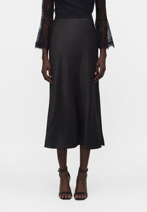 Kobieta ubrana w czarną satynową spódnicę midi, koronkową bluzkę z długim rękawem i czarne wysokie obcasy z paskiem wokół kostki, stojąca na białym tle.