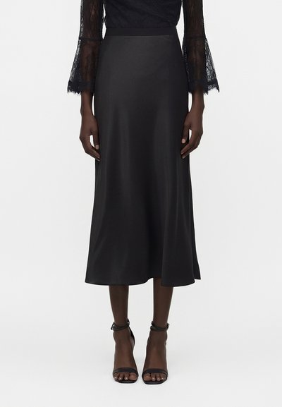 Femme portant une jupe midi en satin noire, un top en dentelle à manches longues et des escarpins noirs à bride à la cheville, debout sur fond blanc.