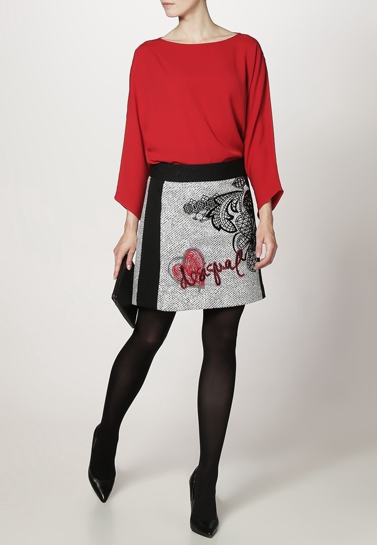 Femme portant un haut rouge à manches longues, une mini-jupe à motif noir et blanc avec une inscription rouge, des collants noirs et des talons pointus noirs, tenant une pochette.