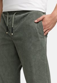 Nahaufnahme einer Person, die olivgrüne Cordhosen mit verstellbarem Bund trägt, mit einer Hand in der vorderen Tasche und einem weißen Hemd.