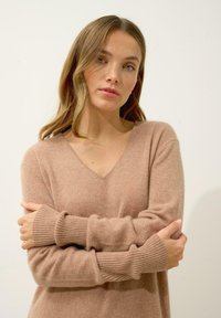 Sweter z dekoltem w kształcie litery V i długim rękawem w miękkim beżowym materiale. Posiada ściągacze na rękawach i luźny krój. Gładka tekstura z minimalnymi detalami.