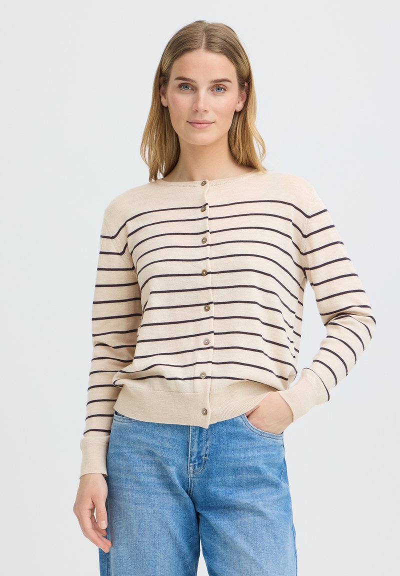 Femme portant un cardigan beige boutonné avec des rayures horizontales noires et un jean bleu, debout avec une main dans la poche devant un fond uni.