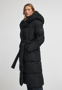 Mujer vestida con un abrigo acolchado negro largo con una gran capucha y un cinturón a la cintura, de pie contra un fondo sencillo.
