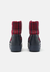 Par de botas de invierno de cuadros azul marino y rojo con parte superior de tela acolchada y suelas de goma gruesas vistas desde atrás sobre fondo blanco.