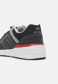 Sportschuhe mit einem dunkelgrauen und marineblauen Textilobermaterial, einer weißen und roten gepolsterten Sohle sowie einem Logo auf der Fersenlasche. Verstärkte Akzente und Mesh-Details.