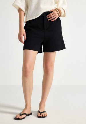 Kvinde iført sorte teksturerede shorts, en løs cremefarvet sweater, træ-armbånd og sorte flip-flops, der står på en hvid gulv.