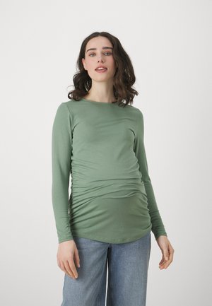 Long-sleeve top in soft green fabric with a round neckline, featuring gathered sides and a fitted silhouette. Worn with light blue jeans.
