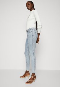 Jeans skinny de color azul claro con un patrón texturizado, que cuentan con una cintura alta y lazos para cinturón, combinados con tacones nude con tiras.