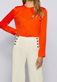 Klart orange långärmad blus med plisserad halsringning. Stylar med högmidjade vita byxor som har guldknappdetaljer. Slät textur.