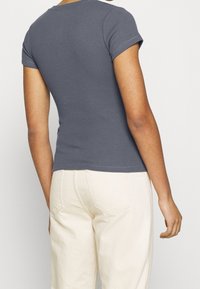 Grå ribbade t-shirt med korta ärmar, figurnära design. Bakvyn visar en rundad halsringning och subtila sömnadsdetaljer. Matchad med beiga byxor.