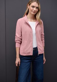 Unge kvinde iført en lys pink hættetrøje med lynlås, hvid skjorte, blå jeans, som holder en brun rem mod en mørk grå baggrund.