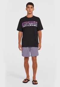 Black t-shirt with bold, multicolored "O'Neill" logo; paired with light purple shorts; model wearing black flip-flops.