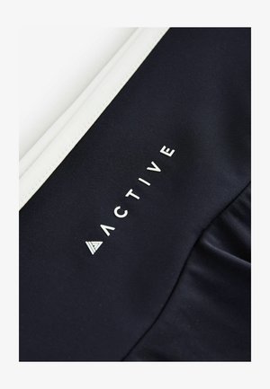 Top de îmbrăcăminte sportivă de culoare bleumarin, cu un finisaj alb, având cuvântul "ACTIVE" tipărit cu litere albe, îndrăznețe, realizat dintr-un material neted și elastic.