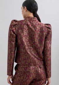 Blazer burdeos con hombros abullonados y patrón abstracto dorado. Textura suave, diseño ajustado y detalle de espalda entallada.