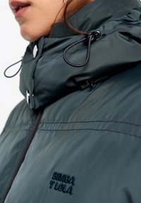 Ciemnozielona kurtka z kapturem o gładkiej, błyszczącej fakturze. Posiada regulowane sznurki, zamek błyskawiczny oraz haftowane logo marki.