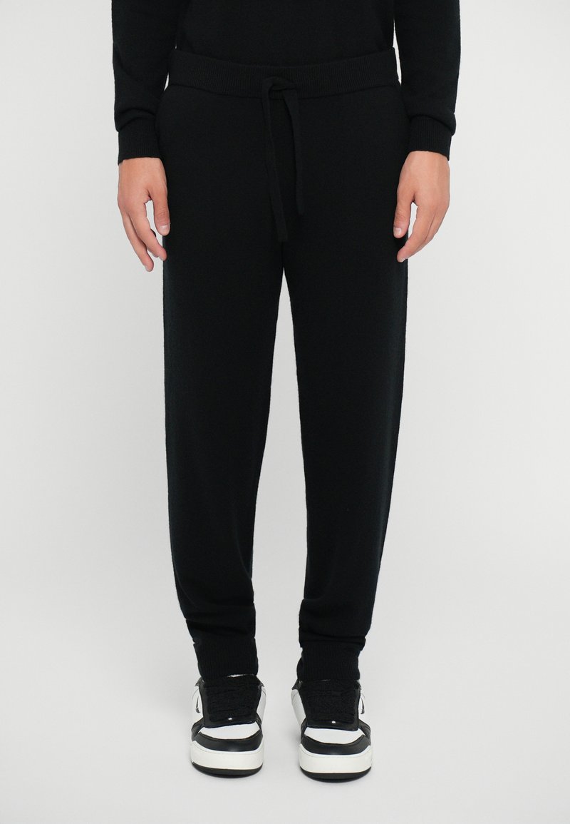Black knitted jogger pants with an elastic waistband and drawstring. Features ribbed cuffs and a relaxed fit. Worn with black and white sneakers.