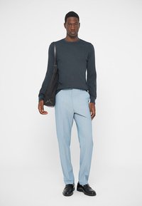 Maglione a coste blu scuro, pantaloni a righe azzurre chiaro, scarpe di pelle nere e una borsa a tracolla nera. Modella in piedi su uno sfondo bianco.