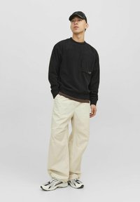 Čierna mikina so predným vreckom, béžové široké nohavice a čierno-biele tenisky. Model stojí na jednoduchom pozadí.