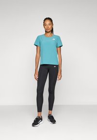 Turkis trænings-T-shirt med korte ærmer og perforeret struktur, parret med sorte leggings og sorte træningssko.
