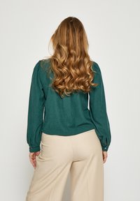 Žalvarinio mėlynumo megzta palaidinė su pūstomis rankovėmis, sagomis uždarytomis rankogaliais ir apvaliu apvadu, derinama su šviesiai beige plačių kojų kelnėmis.