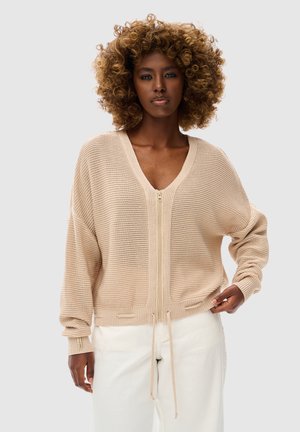 Kobieta z kręconymi włosami, ubrana w beżowy sweter na zamek i białe spodnie, stojąca na jednolitym jasno szarym tle.