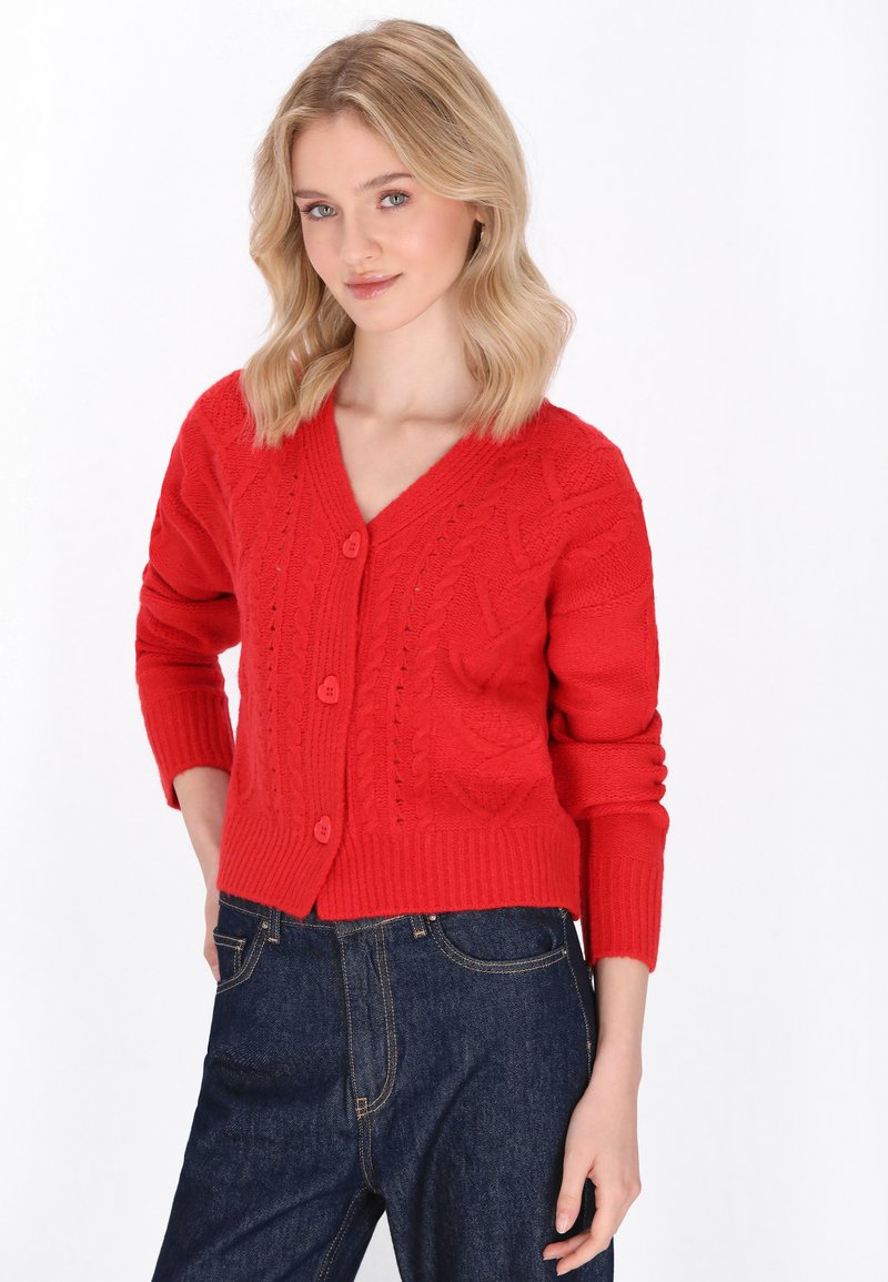 Roter gestrickter Cardigan mit V-Ausschnitt, der strukturierte Muster und drei Knöpfe aufweist, ergänzt durch gerippte Bündchen und Saum. Kombiniert mit dunklen Jeans.