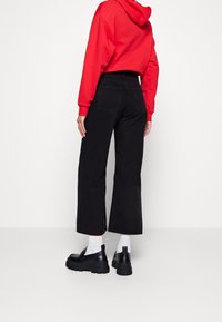 Svarta vida jeans med bakfickor, tillsammans med en röd hoodie. Modellen bär svarta skor med tjocka sulor och vita strumpor.