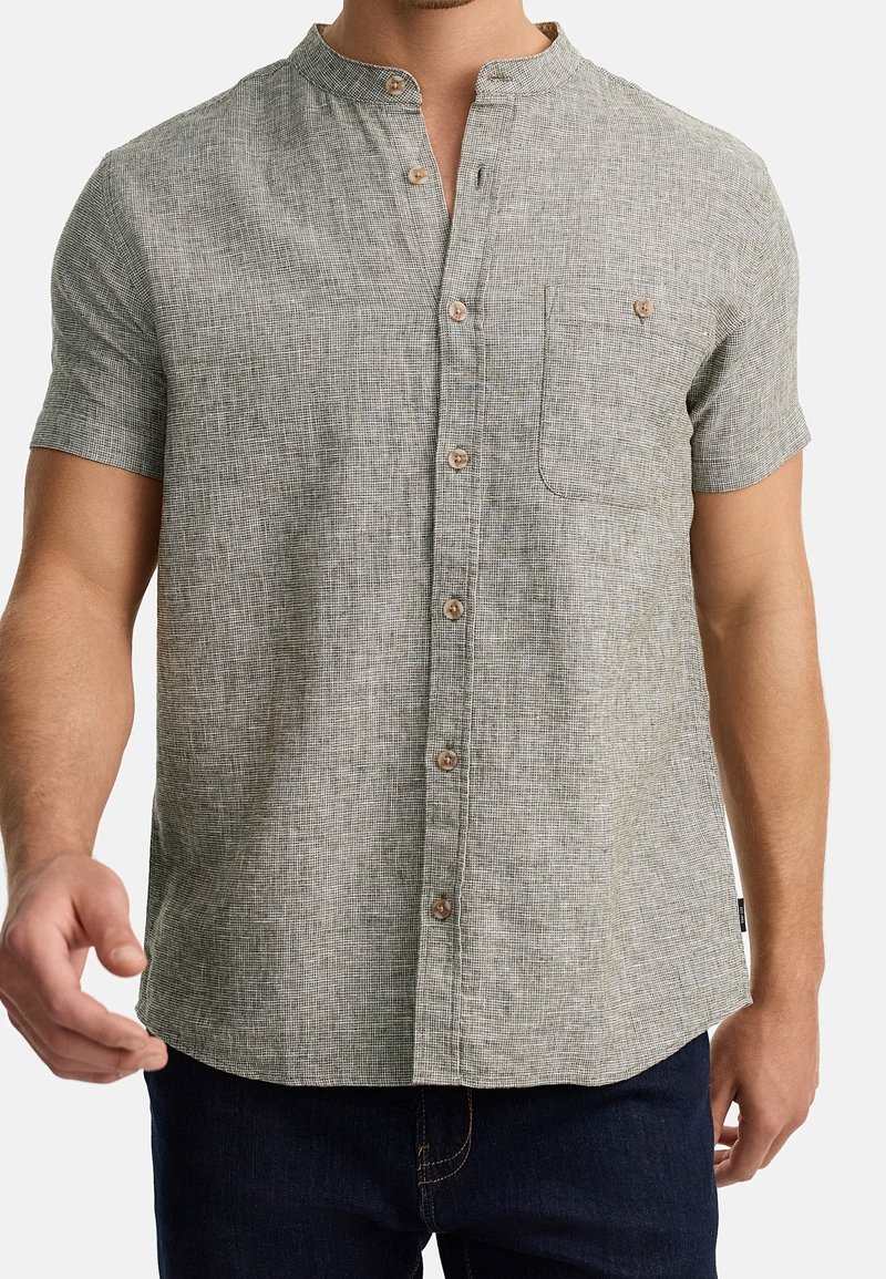 Homme portant une chemise texturée à manches courtes, gris clair, avec une patte de boutonnage devant et un col mao, associée à un jean bleu foncé.