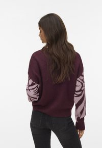 Vrouw met lang bruin haar draagt een paarse trui met roze bloemenpatronen op de mouwen en zwarte jeans, staand tegen een witte achtergrond.