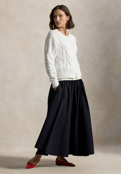 Γυναίκα που φοράει λευκό πλεκτό ζακέτα με κουμπιά, μακριά μαύρη φούστα με τσέπες, μπλε σκουλαρίκια κρεμαστά και κόκκινα μυτερά παπούτσια, στέκεται πλάγια.