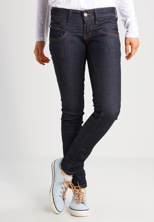 Jambes portant un jean skinny bleu foncé et des baskets bleu clair avec des lacets beige, debout avec un pied croisé sur l'autre.