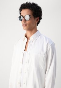 Occhiali da sole rotondi con lenti scure, montatura in metallo argentato e riflesso visibile. Indossati da una persona con una camicia bianca con bottoni.