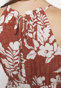 Gros plan d'une personne portant une robe sans manches rouille et blanche à motifs floraux avec un nœud noué dans le dos au niveau de l'encolure.
