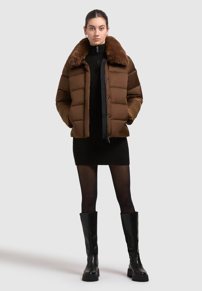 Brązowa kurtka puchowa z kołnierzem z sztucznego futra, zapinana na guziki, z pikowanym wzorem. Połączona z czarną sukienką z prążkowanej tkaniny i czarnymi kozakami do kolan.
